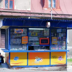 Kiosk Lotto z ofertą zdrapek. Czy warto kupować zdrapki na stacjach benzynowych czy lepiej w kolekturze? Dwoje młodych ludzi siedzi obok.