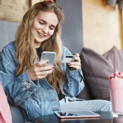 Uśmiechnięta kobieta w dżinsowej kurtce trzyma telefon i kartę płatniczą.