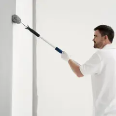 Mężczyzna w białej koszulce maluje ścianę wałkiem. Odkrywamy sekrety produkcji farby do zdrapywania, której nie da się prześwietlić.