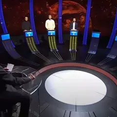 Trzech uczestników teleturnieju stoi przy swoich stanowiskach, a prowadzący siedzi przodem do nich.