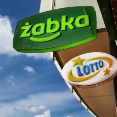 Zielona tablica "Żabka" i biała z napisem "LOTTO" wiszą na tle błękitnego nieba z chmurami.