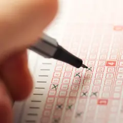Dłoń z długopisem zaznacza liczby na kuponie Lotto.