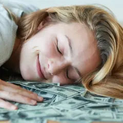 Uśmiechnięta kobieta śpi na stosie banknotów dolarowych. Marzy o tym, czy polskie gwiazdy rzeczywiście biorą udział w międzynarodowych kumulacjach.