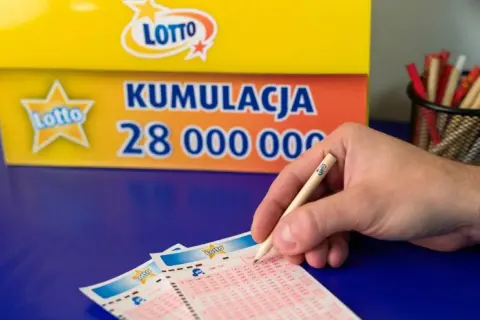 Ręka z ołówkiem Lotto nad kuponami. Tło: baner "Kumulacja 28 000 000". Czy wygrana w małej miejscowości sprawia, że sąsiedzi stają się wrogami?