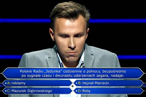Młody mężczyzna w szarym garniturze zastanawia się nad pytaniem z teleturnieju. Jakie pytania zadaje psycholog zwycięzcom lotto w poradniku milionera?