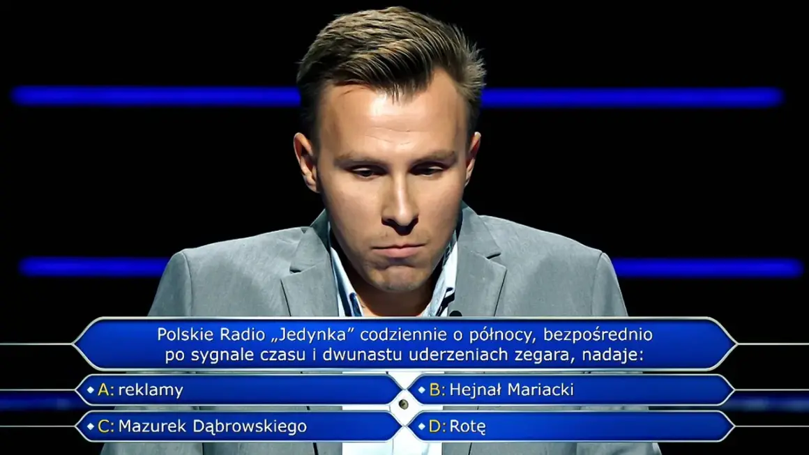 Młody mężczyzna w szarym garniturze zastanawia się nad pytaniem z teleturnieju. Jakie pytania zadaje psycholog zwycięzcom lotto w poradniku milionera?