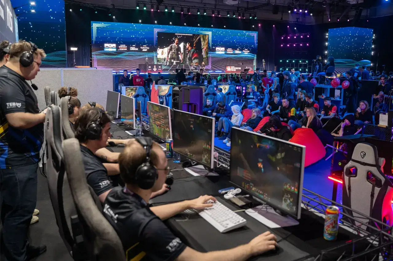 Uczestnicy turnieju esportowego siedzą przy komputerach, gotowi do rywalizacji. Aby wziąć udział w eliminacjach, muszą spełnić określone wymagania techniczne, takie jak posiadanie odpowiedniego sprzętu i stabilnego połączenia internetowego.