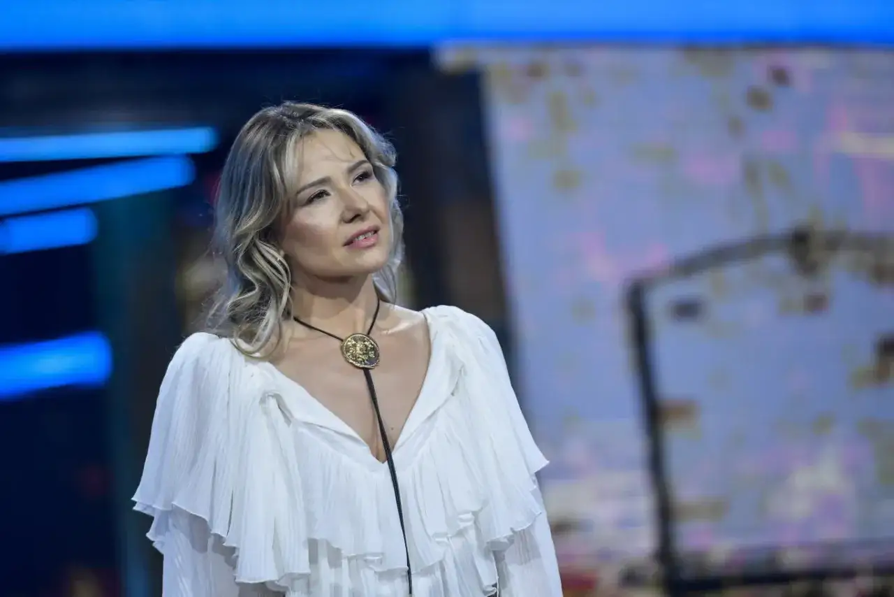 Wokalistka w białej sukni z falbanami i złotą broszką, zastanawia się, dlaczego popularna wokalistka została odsunięta od śpiewania w programie.