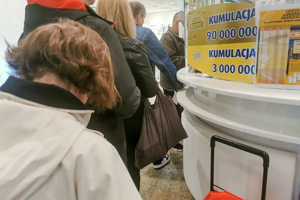Ludzie stoją w kolejce do punktu Lotto, gdzie widnieje informacja o kumulacji. To może być jeden z prawdziwych powodów dla których zwycięzcy loterii ukrywają swoją tożsamość.
