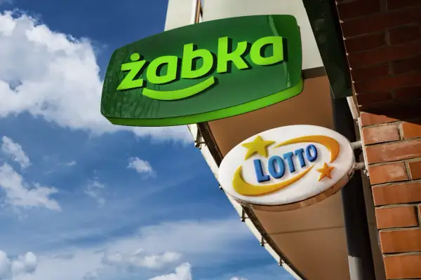 Zielona tablica "Żabka" i biała z napisem "LOTTO" wiszą na tle błękitnego nieba z chmurami.